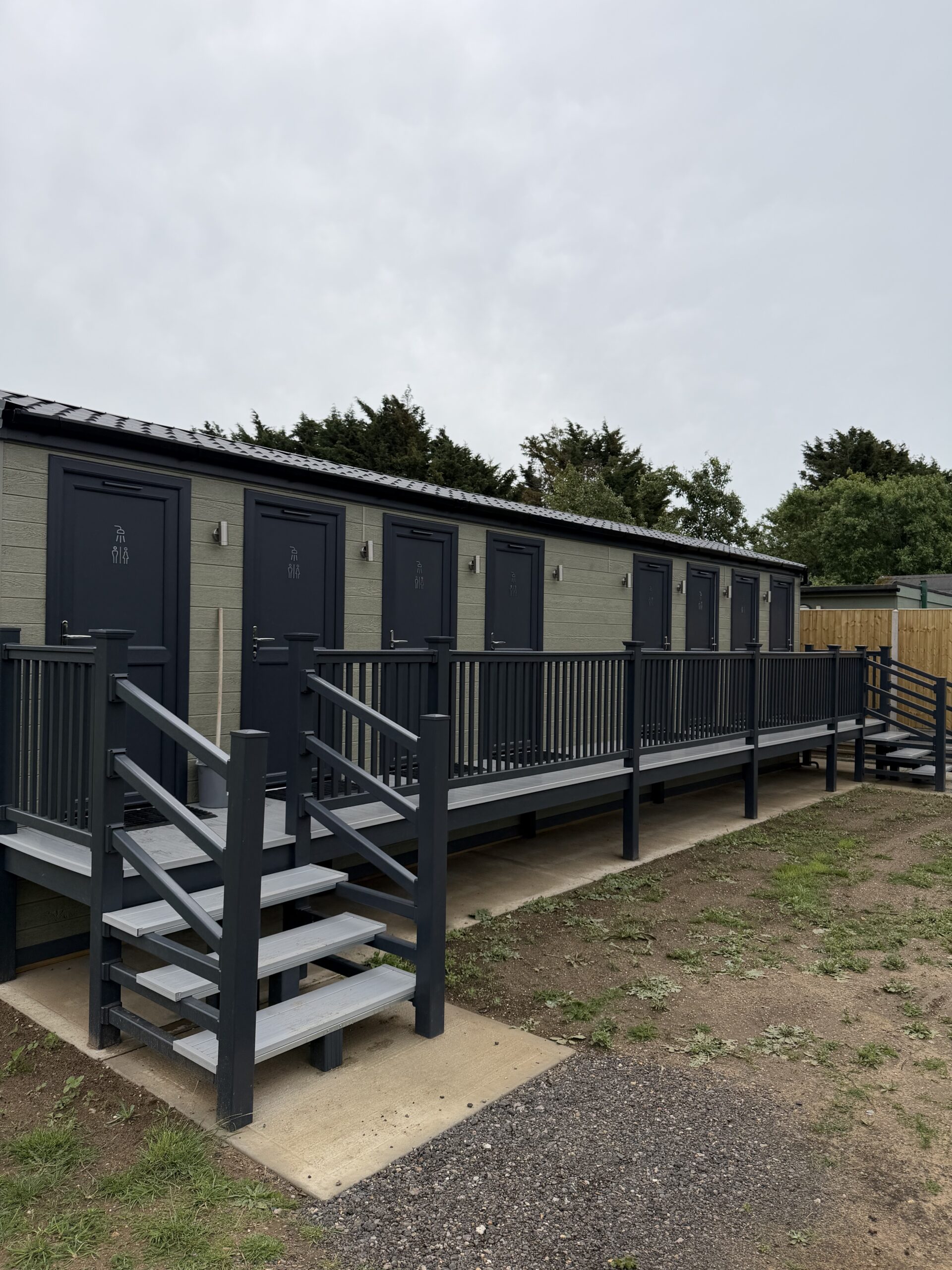 Low Farm 1 | Arkem Leisure Ltd Row of dark blue portable toilets with stairs and railings along a wooden deck, outdoors under a cloudy sky, with trees in the background.